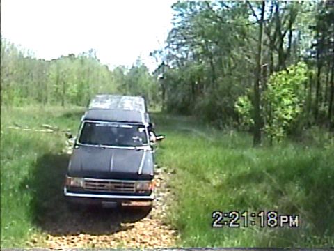 1988 Ford Bronco - Van driving on a dirt road surrounded by trees and grass.