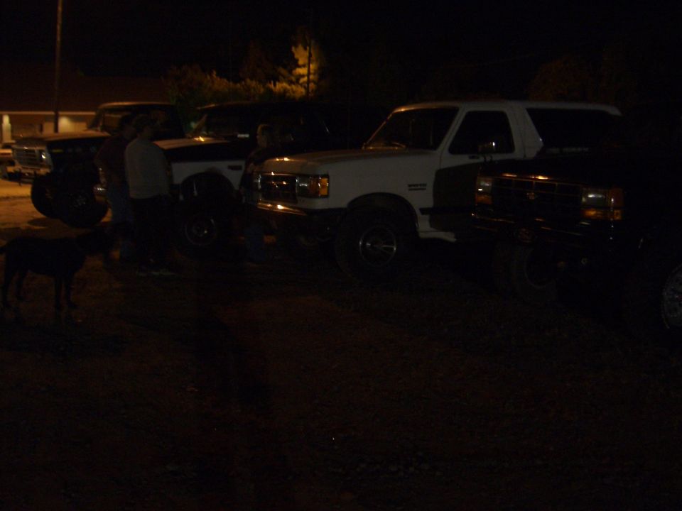 White Ford Bronco parked at night beside other trucks with people and a dog on gravel