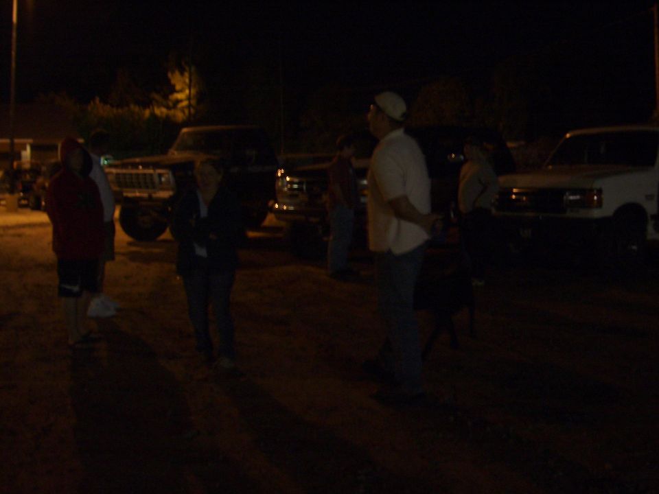 People standing in a dark dirt lot at night beside parked trucks and a white SUV