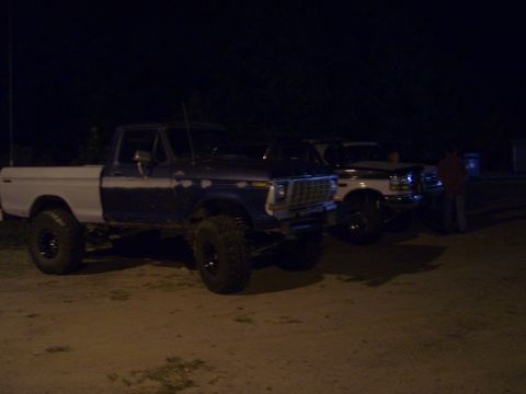 1996 Ford Bronco - Two lifted Ford F-Series pickup trucks are parked side-by-side at night, with one truck showing primer on its body panels.