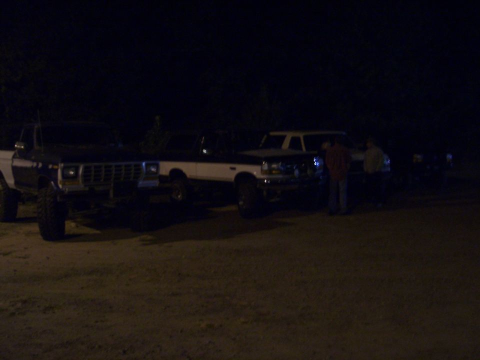 Row of lifted Ford trucks parked on dirt lot at night with people nearby