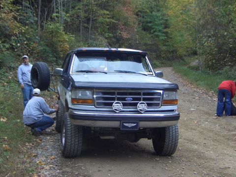 1996 Ford Bronco - A Ford Bronco is parked on a dirt road in a wooded area, with people attending to it and a spare tire mounted externally.