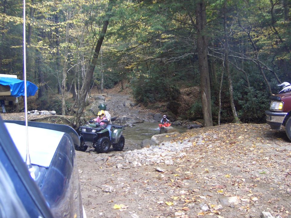 ATV riders at a rocky creek crossing with vehicles parked on a wooded trail