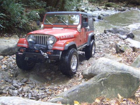 1996 Ford Bronco - A red Jeep CJ-7 Laredo navigates a rocky stream bed surrounded by trees and foliage.