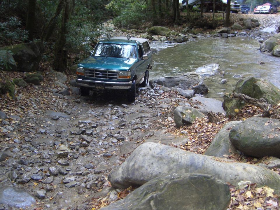 Teal Ford Bronco with white top crossing a rocky creek in the woods