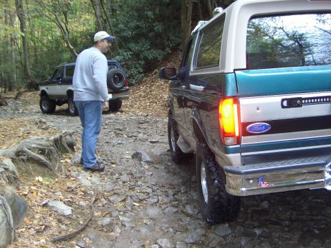 1996 Ford Bronco - Two Ford Broncos navigate a rough, off-road path surrounded by trees and fallen leaves.