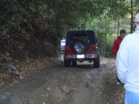 1996 Ford Bronco - A red Jeep Wrangler navigates a muddy trail surrounded by trees, with other vehicles and people present.