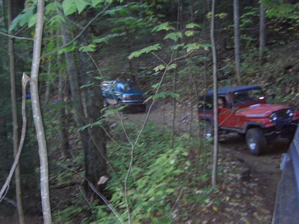 Red Jeep Wrangler and blue truck on a wooded off-road dirt trail