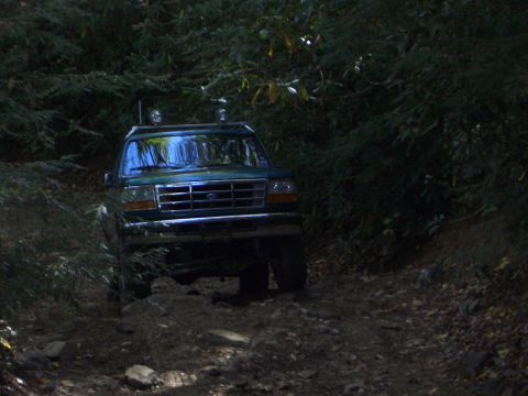1996 Ford Bronco - A green SUV navigates a rough, wooded trail, its headlights reflecting the dim forest light.