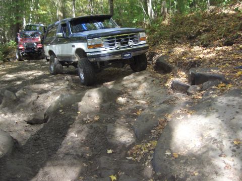 1996 Ford Bronco - Two SUVs navigate a challenging rocky path in a wooded area during autumn.