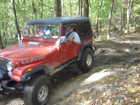 1996 Ford Bronco - A red Jeep CJ-7 Laredo navigates a rocky off-road trail surrounded by trees.