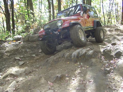 1996 Ford Bronco - A red Jeep Wrangler navigates a challenging rocky path through a wooded area.