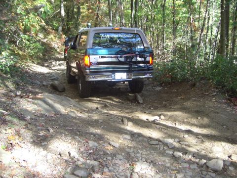 1996 Ford Bronco - A 1996 Ford Bronco navigates a challenging rocky path through a wooded area. Another vehicle is ahead on the trail.