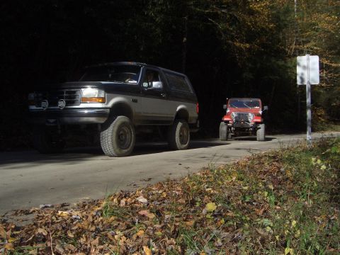 1996 Ford Bronco - A 1996 Ford Bronco and a red Jeep drive down a road lined with autumn foliage.