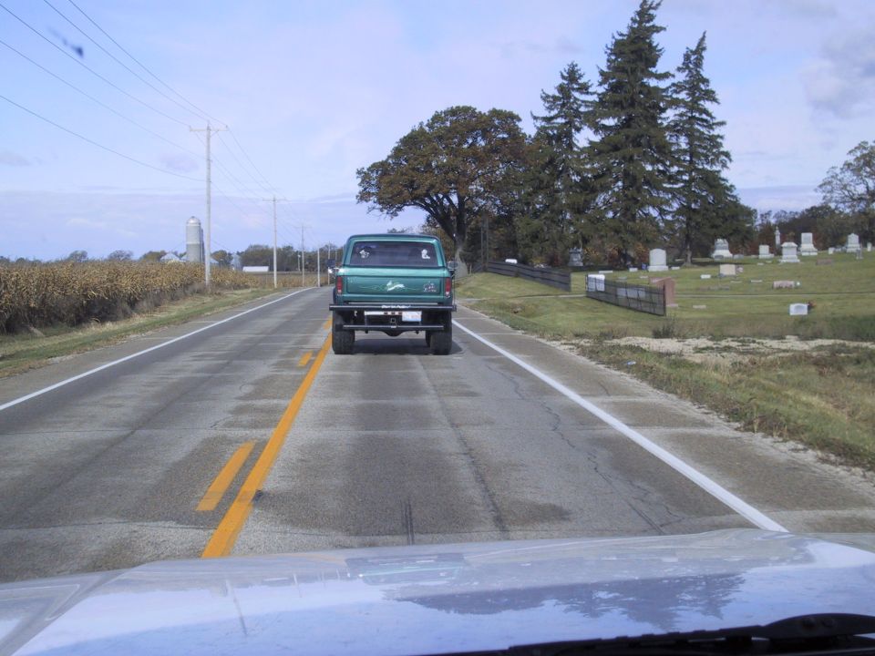 Green pickup truck seen from behind on a two-lane road, with a cemetery on the right