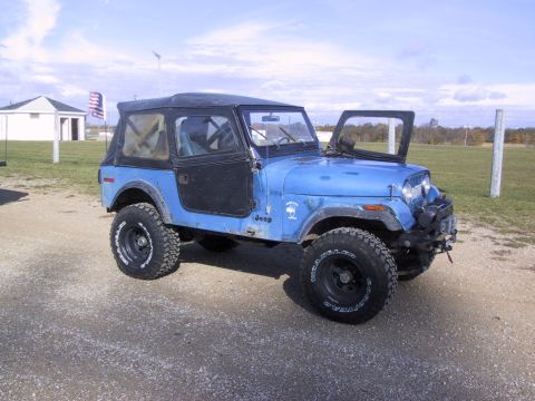 1993 Ford Bronco - A blue Jeep CJ-7 with its doors off sits parked on a gravel surface under a cloudy sky.