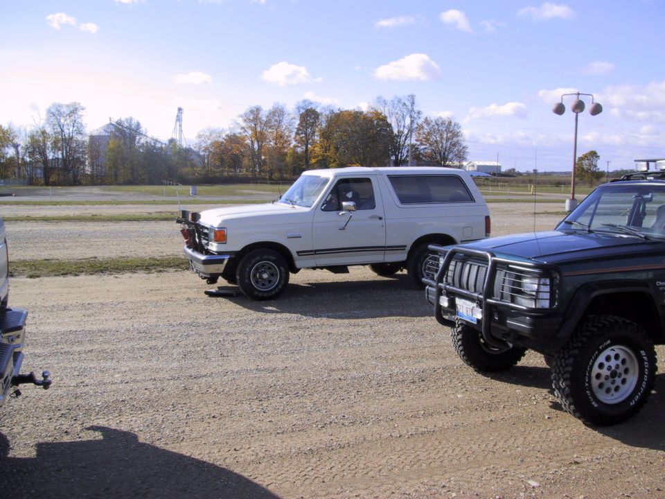 White 1993 Ford Bronco with aftermarket lights parked next to a dark SUV