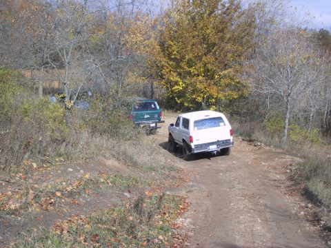 1993 Ford Bronco - Two SUVs drive on a dirt road surrounded by trees with autumn foliage.