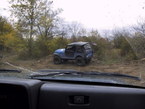 1993 Ford Bronco - A blue Jeep Wrangler navigates a dirt path surrounded by trees and foliage.