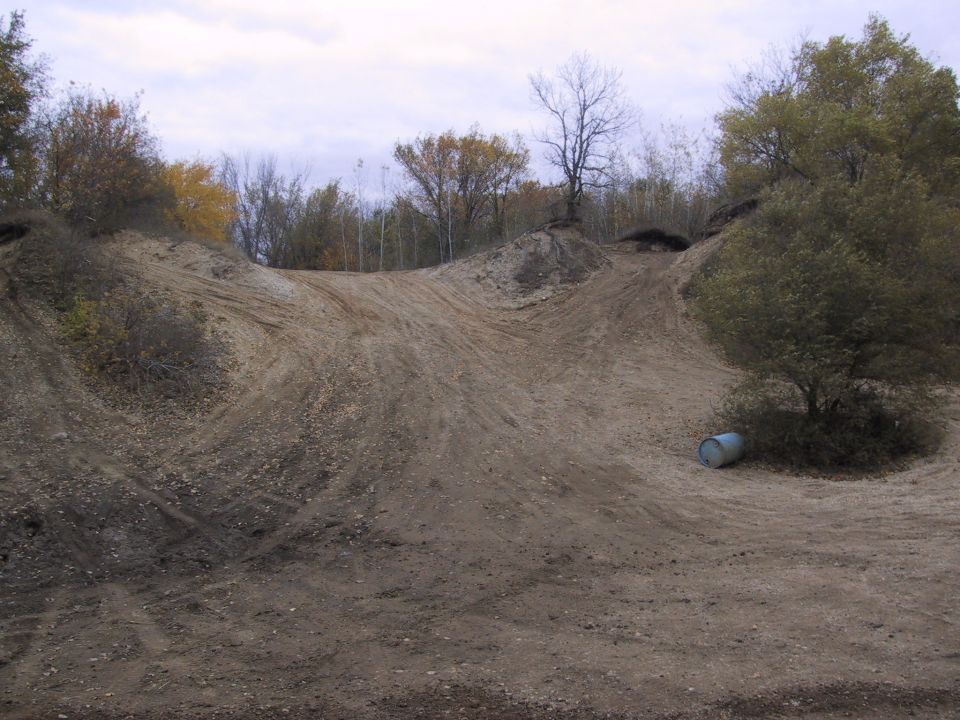 Dirt track with tire marks and a barrel under a tree