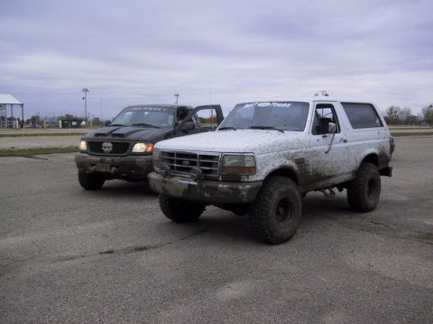 1993 Ford Bronco - Two off-road vehicles, a white Bronco and a black pickup truck, are parked side-by-side after a muddy excursion.