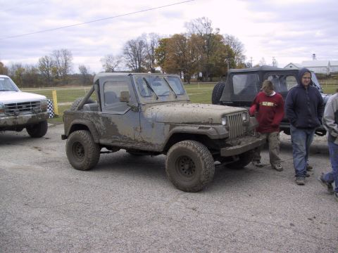 1993 Ford Bronco - A muddy Jeep Wrangler sits parked on a gravel lot with people standing nearby. Another vehicle is visible to the left.