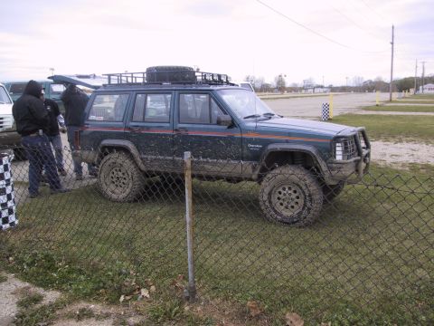 1993 Ford Bronco - A mud-covered Jeep Cherokee XJ sits on grass, with people standing nearby and a race track in the background.