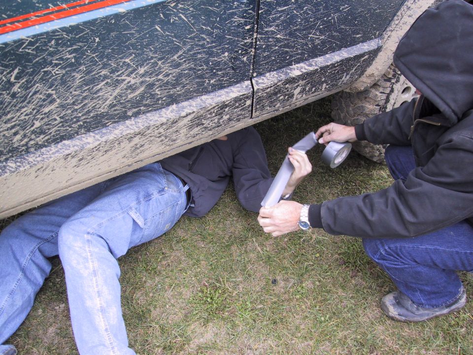 Mud-splattered dark green SUV side with two people working underneath on exhaust pipe with duct tape
