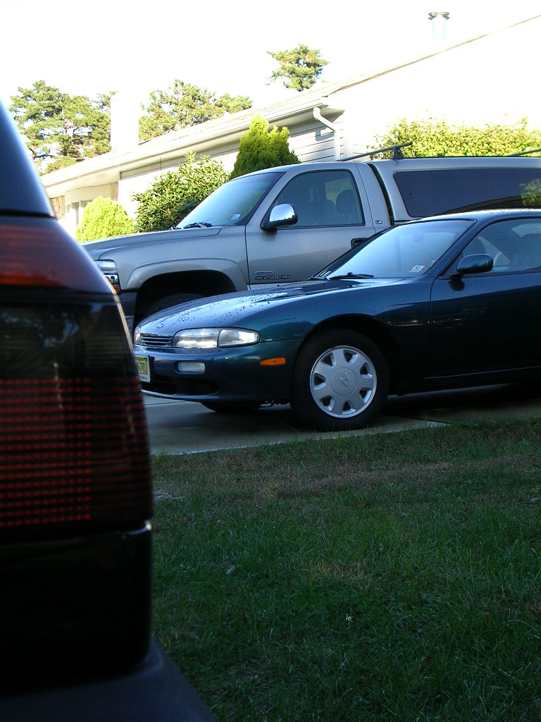 Dark green coupe with silver hubcaps, 3q-front view, parked next to a silver pickup truck
