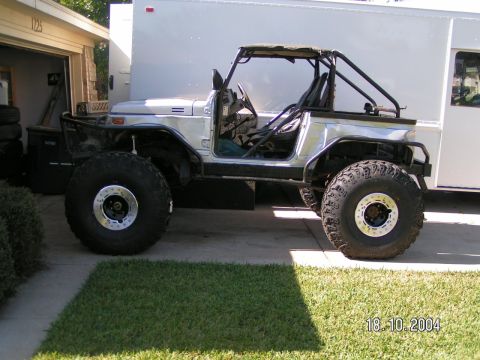 1978 Ford Bronco - A modified off-road vehicle with a roll cage and oversized tires sits in a driveway next to a garage and a large white trailer.