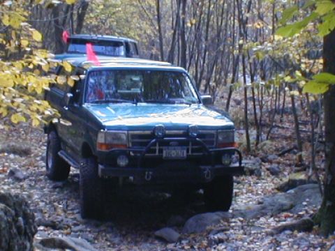 1979 Ford Bronco II - Two Ford Broncos navigate a wooded trail with fallen leaves and rocks.