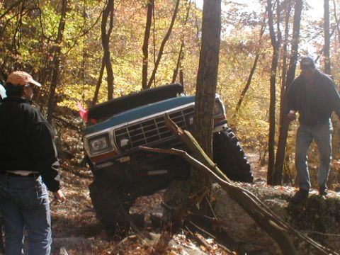 1979 Ford Bronco II - An off-road SUV is stuck on a rocky trail, leaning against a tree with two men observing the situation.