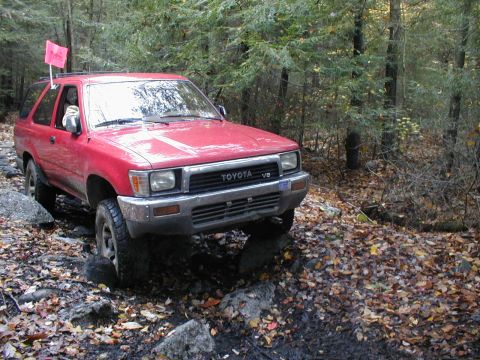 1979 Ford Bronco II - This red Toyota 4Runner is shown off-roading through a wooded area with fallen leaves on the ground.