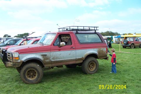 1996 Ford Bronco - A muddy Ford Bronco is parked on grass, with other vehicles and tents in the background.