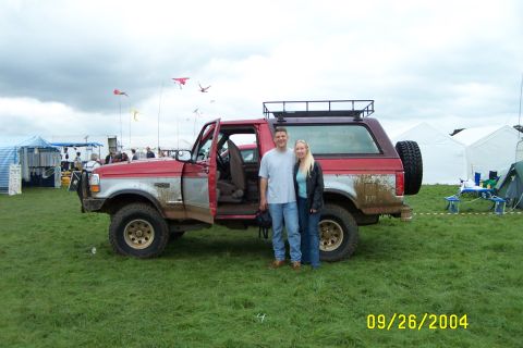 1996 Ford Bronco - A couple poses with a mud-splattered Ford Bronco at an outdoor event with tents and kites in the background.