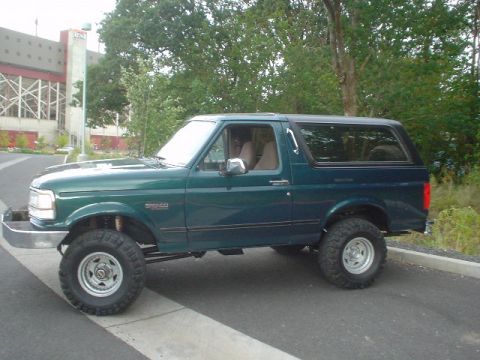 1993 Ford Bronco - A lifted green Ford Bronco is parked on a paved surface with trees and a building in the background.