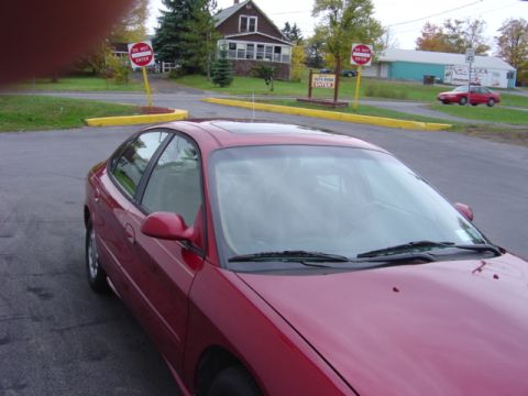 1998 Ford Taurus - A red 1998 Ford Taurus sedan is parked on an asphalt lot near buildings and signs.