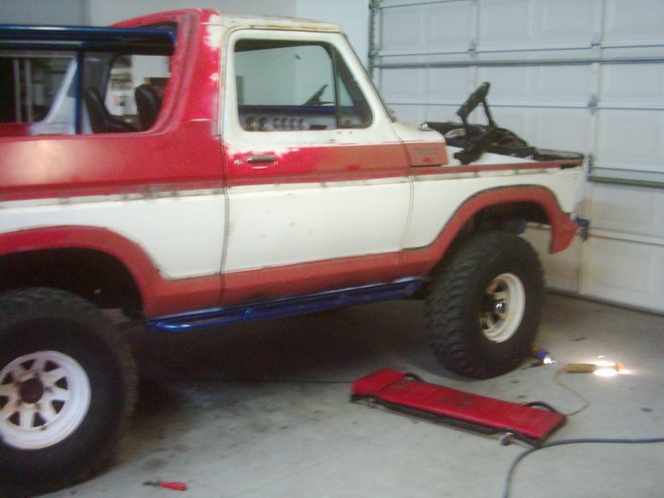1978 Ford Bronco with red and white paint, blue side steps, and roll cage, parked in a garage