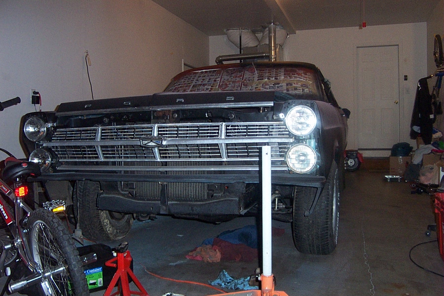 Front of dark classic Ford on jack stand in garage, bumper off, chrome grille exposed