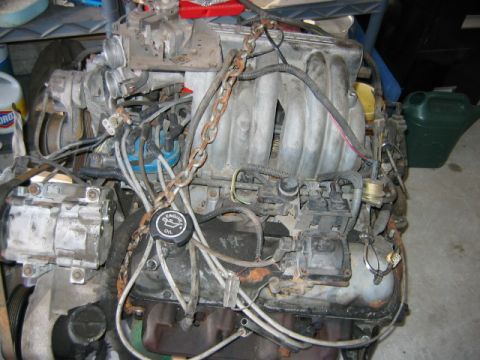 1992 Ford Bronco - A V-engine removed from the vehicle, viewed from above, showing an aluminum intake manifold, a black oil-filler cap, a belt-driven A/C comp…