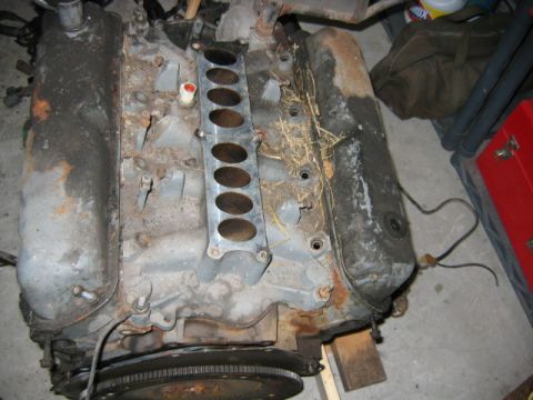 1992 Ford Bronco - A dusty V-configuration engine block sitting on a workbench with an intake manifold featuring multiple round ports centered on top; surface…
