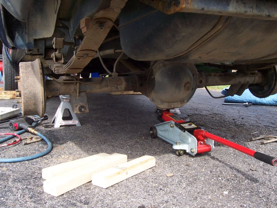 Vehicle's rear axle assembly supported by jack stands, with a floor jack positioned under the differential. Leaf springs and fuel tank are visible