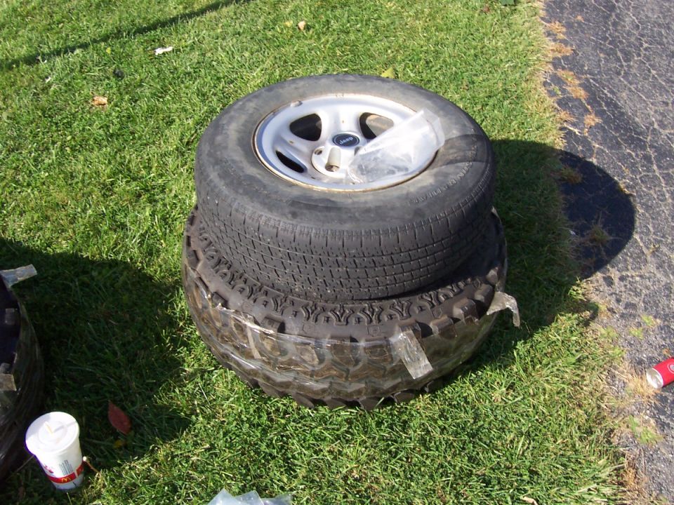 Street tire on white Jeep wheel stacked atop a larger mud-terrain tire on grass