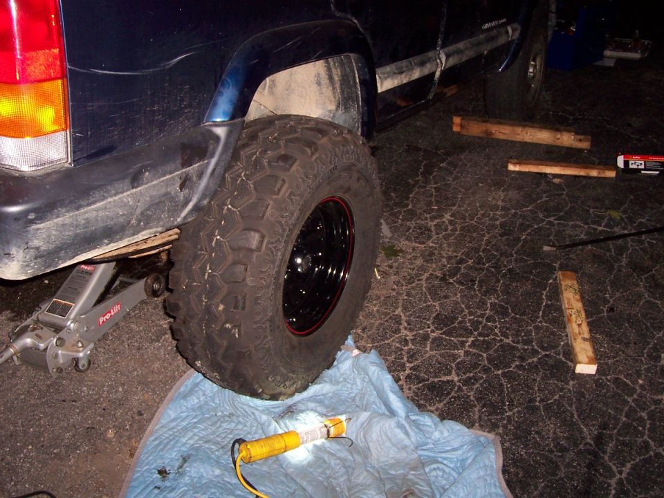Side view of a 2000 Jeep Cherokee with a jack under the rear axle and a large tire