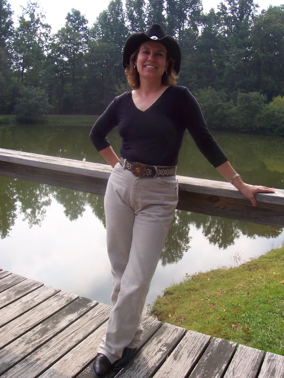 Woman in black cowboy hat and top leaning against a wooden rail by a pond