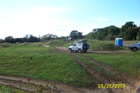 1996 Ford Bronco - Two off-road vehicles are seen on a dirt track at a grassy off-road park under a clear blue sky.