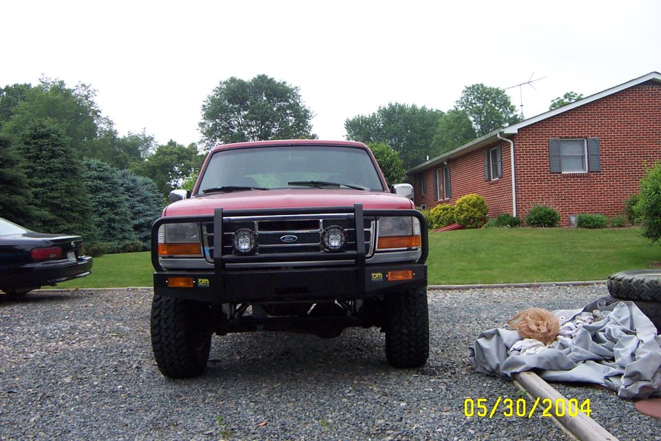 Red lifted Ford truck with black brush guard and off-road tires, front view on gravel