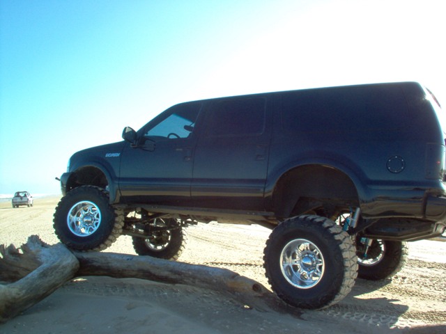 Lifted black Ford Excursion SUV with big off-road tires and chrome wheels on a sandy beach, side view