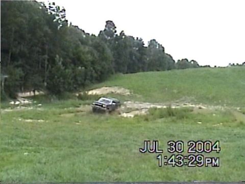 1988 Ford Bronco - A dark SUV navigates a muddy terrain on a grassy hill, with trees lining the horizon.