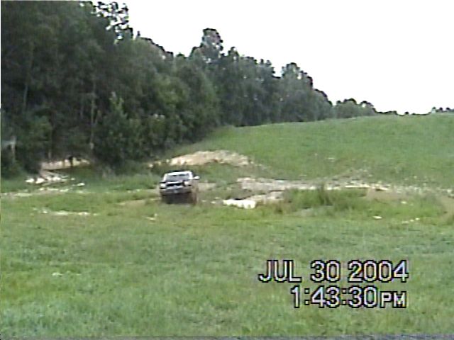 Truck driving through a muddy, grassy field with trees in the background. Timestamp overlay reads JUL 30 2004 1:43:30 PM